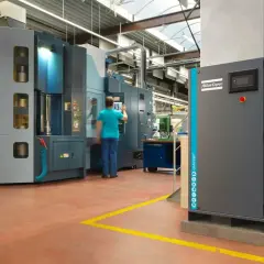 En una fábrica, un operario interactúa con una máquina industrial. A la derecha, un compresor Atlas Copco, que puede ser con aceite o sin aceite, espera su turno.
